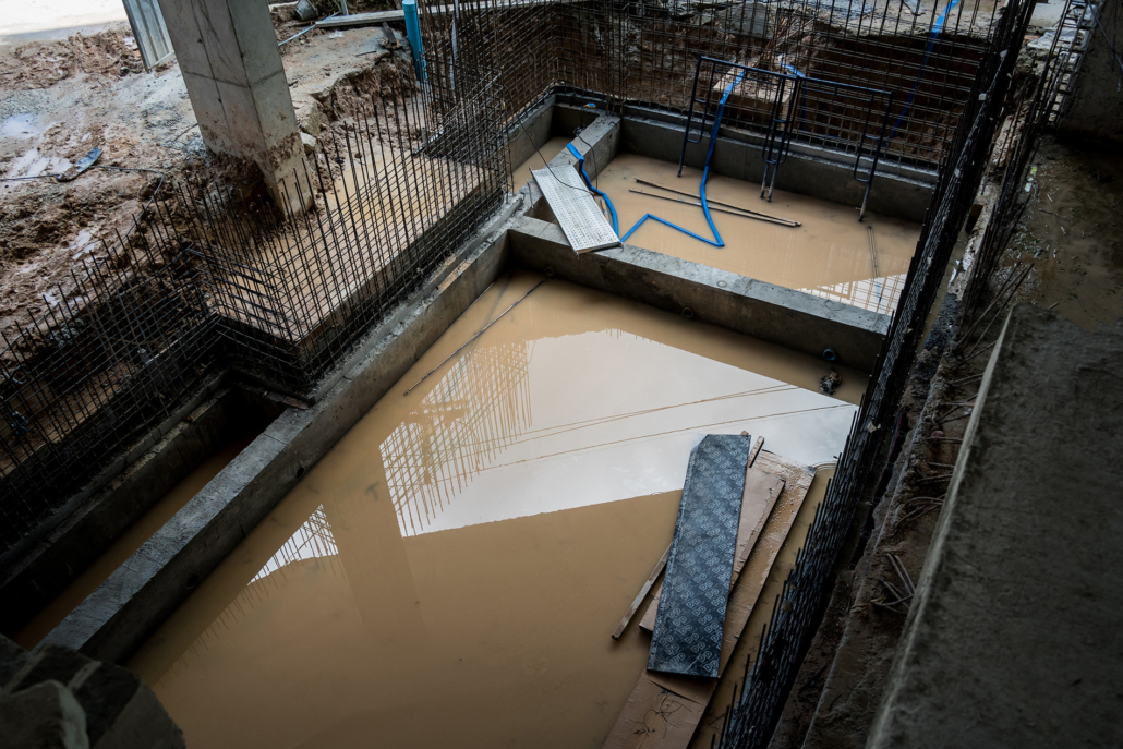 Partially constructed concrete foundation filled with muddy water, metal reinforcement bars, and construction materials scattered around the site. Reflections are visible on the water surface.